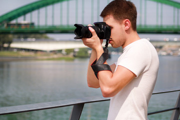 Fototapeta na wymiar Professional photographer examining scenery and taking pictures of environment, architecture, urban elements, river, old green bridge. Tourist and traveler is making landscape photography with camera.