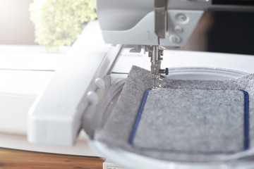 detail view on a modern computerised sewing machine with embroidery unit in bright light stitching a blue frame on grey felt in a friendly work environment with flowers