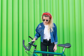 Young woman in stylish clothes stands with a bicycle on the background of a green wall. Teen girl walking with a bike. Woman is a cyclist. Copyspace.