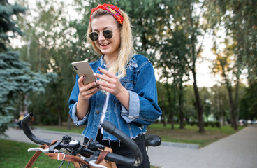 Happy, stylish girl sitting on a bike in the park, using a smartphone and smiling. Walk on the bike in the park. Hipster girl with a bike and a smartphone in the park