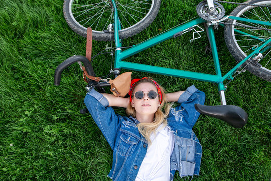 Portrait of an attractive stylish girl lying on the grass with a bike and looking up. Fashionable woman dressed in a denim jacket and sunglasses lays on the grass bike. Copyspace