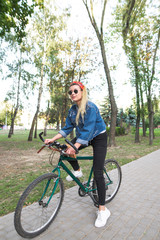 Obraz premium Attractive girl in stylish clothes sitting on a bike in the park. Teenage girl in sunglasses and a denim jacket is in a park with a bicycle. Walk on the bike.