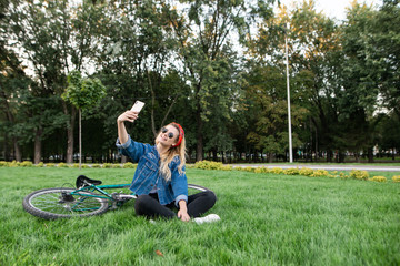 Young girl sits in the grass in park and takes selfie on a smartphone. Happy beautiful girl in stylish clothes and sunglasses sits on the grass the park and takes selfie on the background bike.