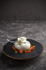 Whole tied Italian cheese burrata on small wooden plate served with fresh tomatoes and basil on dark textured background