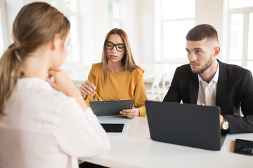 Young business man with laptop and business woman in eyeglasses with folder and pencil in hands thoughtfully talking with applicant about work. Young employers spending job interview in office
