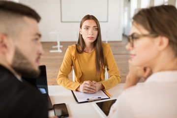 Young pensive woman in blouse dreamily looking at employers while spending time in modern office. Female applicant waiting results of job interview