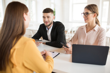 Smiling business man with tablet and business woman with mobile phone in hands while joyfully talking with applicant on work. Young employers spending job interview in modern office