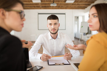 Young man in white shirt thoughtfully looking at employers while spending time in modern office. Male applicant holding pencil waiting results of job interview