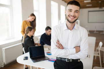 Young smiling man with beard in white shirt happily looking in camera with folded hands while spending time in office with colleagues on background