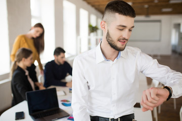 Young man with beard in white shirt  looking on handwatch while spending time in office with colleagues on background