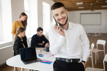 Young smiling man with beard in white shirt joyfully looking in camera talking on cellphone while spending time in office with colleagues on background