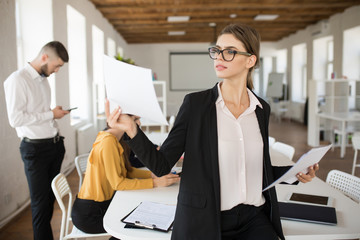 Young business woman in eyeglasses and shirt thoughtfully watching documents while working in office with colleagues on background
