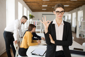Young smiling business woman in eyeglasses and shirt dreamily looking in camera showing ok gesture while spending time in office with colleagues on background