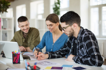 Young man in shirt and eyeglasses thoughtfully working in office with colleagues on background
