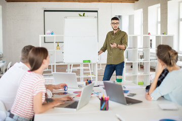 Obraz premium Young man in eyeglasses and shirt standing near board while discussing new project with colleagues at work. Group of people working together in modern office