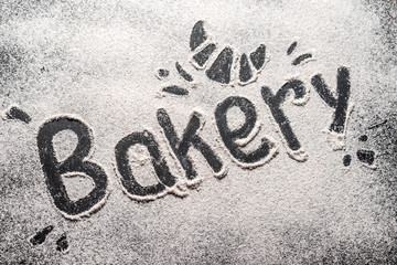 The bakery inscription is made on white wheat flour scattered on a black table.  Top view, space for text. Flour ready to knead dough for baking bread, cake or cake