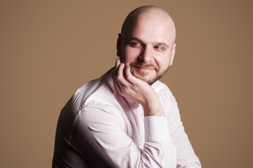 Portrait of successful handsome bearded bald man in light pink shirt and white bow, sitting on chair and looking away with smiley face and pondering. indoor studio shot, isolated on brown background.
