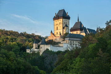 Famous historical medieval castle Karlstejn built by emperor Charles IV., lit by evening sunlight, standing on a green hill covered with trees, Czech Republic, Europe, blue sky