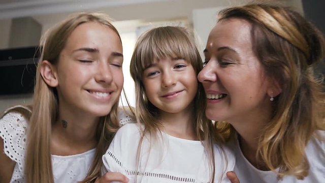 The charming girl with long hair in white dress stands and looks in the camera, mother and older sister fit hug and kisses her at home in the kitchen. Portrait of a lovely family