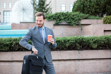 Obraz premium Happy young businessman standing in front of an office building, checking time on his wrist watch. Carrying a briefcase and holding a cup of coffee. Work anywhere concept.
