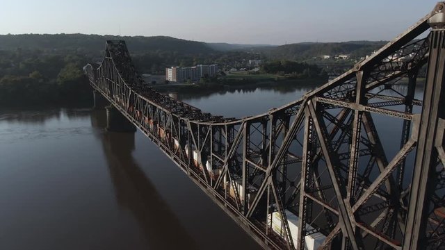A high angle aerial profile view of a cargo train traveling over a railroad bridge spanning the Ohio River. Pittsburgh suburbs.  	