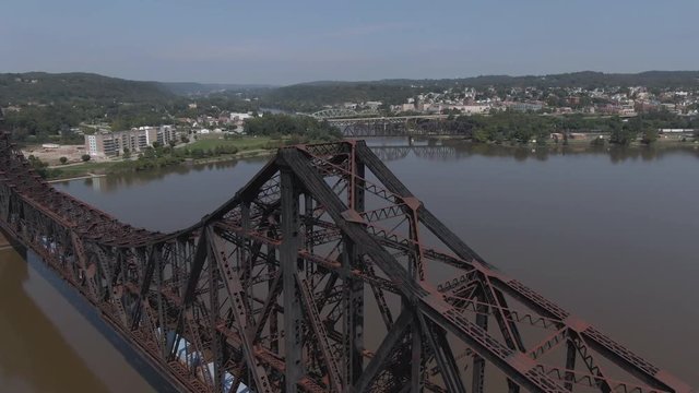 A cinematic aerial orbiting establishing shot of a small western Pennsylvania town on a summer day. Large railroad bridge in the foreground. Pittsburgh suburbs.  	