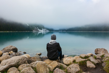 Fototapeta na wymiar Man sitting on the rocks at Lake Louise on a foggy day