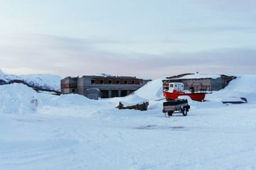 a trailer for the car and a small red boat stand outside in a cold winter day in the snow in the north near an abandoned and destroyed brick building