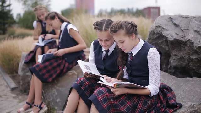 Group of young friends students wearing the same school uniform sitting and talking outdoors while reading a book on campus.