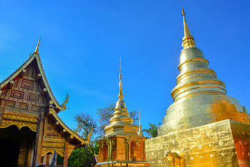 Naklejka premium golden Buddhist temple, shiny golden pagoda and wooden church at Wat Pra Sing with blue sky background, Chiang-mai province northern of Thailand