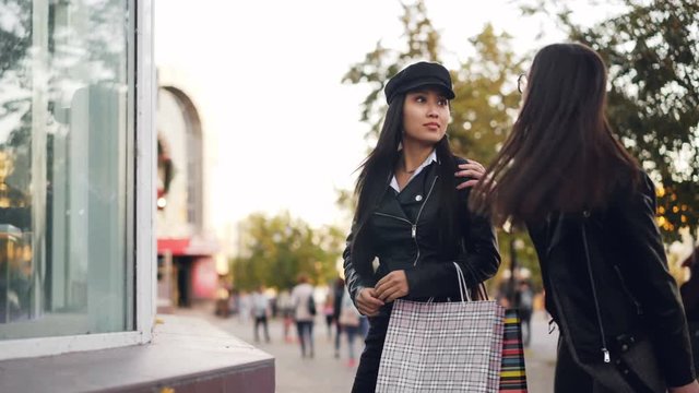 Cute Asian girl is looking at things in shop window then meeting her friend, young women are hugging and talking showing purchases in shopping bags then leaving.