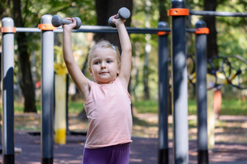 The child high raises dumbbells. A little girl with blond hair goes in for sports.