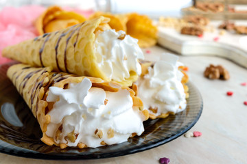 Delicious honeyed waffles, twisted into cones with cream, nuts, chocolate topping on the plate on white wooden background.