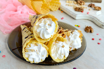 Delicious honeyed waffles, twisted into cones with cream, nuts, chocolate topping on the plate on white wooden background.