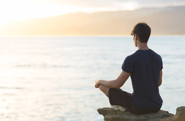 Young man meditating on top ocean cliff during sunset.