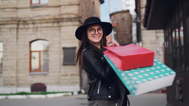 Slow motion portrait of happy young lady with long dark hair wearing glasses and hat walking with shopping bags then turning and looking at camera with glad smile.