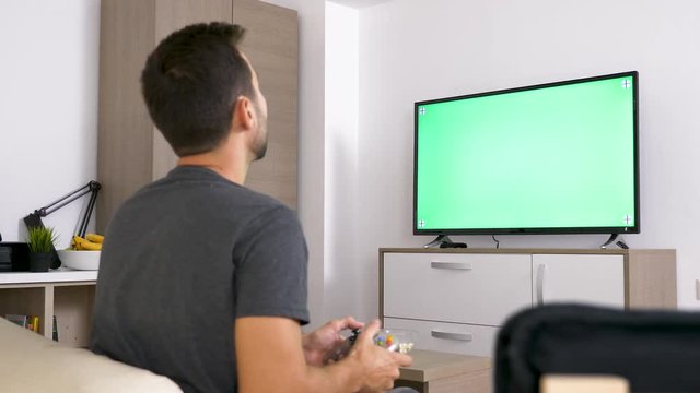 Adult man in front of big green mock-up screen TV playing a video game on the console. Static shot. 4K footage