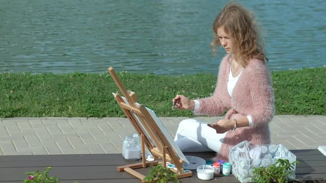 A pretty woman with red hair, paints a picture on canvas, which stands on the easel. The lady is in the open air near the lake of the river, she draws from life