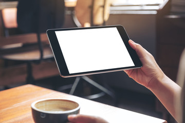 Obraz premium Mockup image of woman's hands holding black tablet pc with blank white screen while drinking coffee on wooden table in cafe