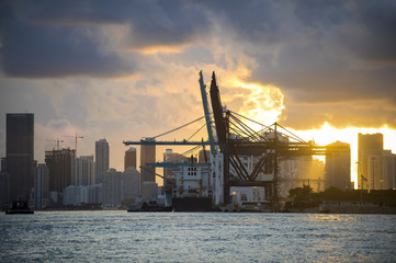 Container ship being unloaded with cranes at a port with city skyline backdrop at sunset