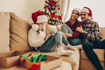 Christmas. Family. Technologies. Little girl is using a digital tablet while her parents are watching her and smiling. At home near the Christmas tree
