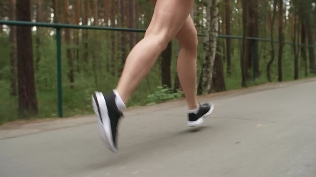 Low-section dolly shot of legs of unrecognizable sportswoman running along road in park