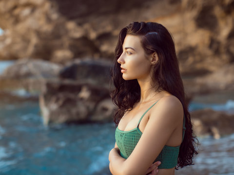 Fashion young girl in bikini outdoors against wild rocky tropical background. Close-up portrait of beautiful sensual brunette mixed race Asian Caucasian woman standing on the beach near rock. Slim