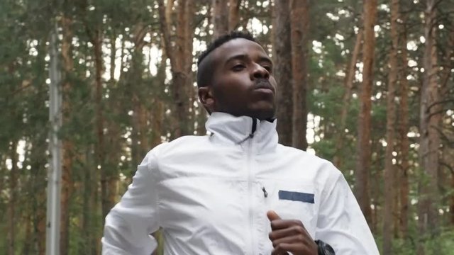 Dolly shot of black man wearing windbreaker running in forest on chilly morning