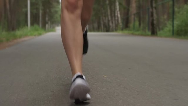Low angle dolly shot with tilt up of sporty young woman with fit body running along road in forest