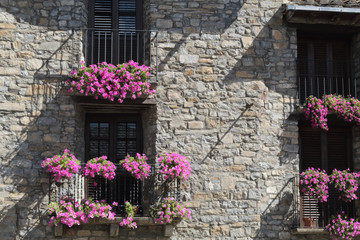 A front view of a stone facade with four black iron railing balconies with violet hanging flower on them in Ainsa, a small rural village in the Spanish Aragonese Pyrenees mountains