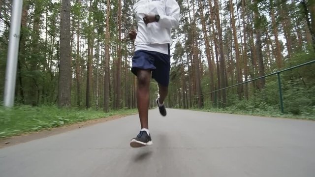 Low angle dolly shot of unrecognizable black man in shorts and windbreaker running along road in forest