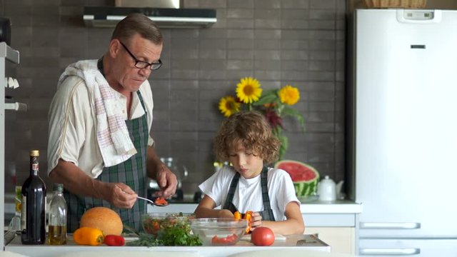 Grandson and grandfather cook together in the kitchen. An elderly man gives to try a salad boy. Healthy lifestyle, modern kitchen interior