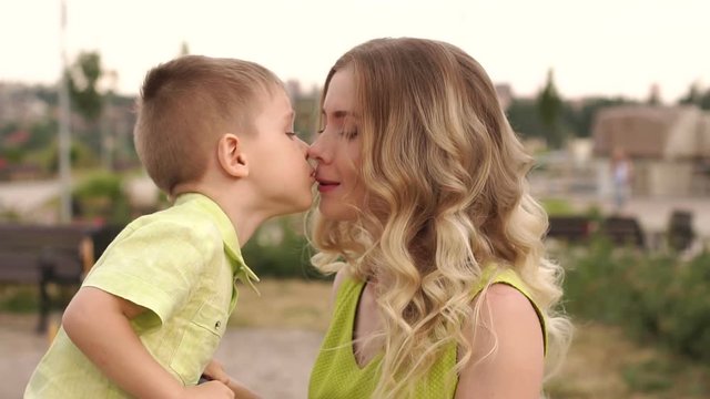 Close-up of a little cute boy kissing his beloved mommy in the park in the summer. Portrait of a happy mother and her little son in a playground.