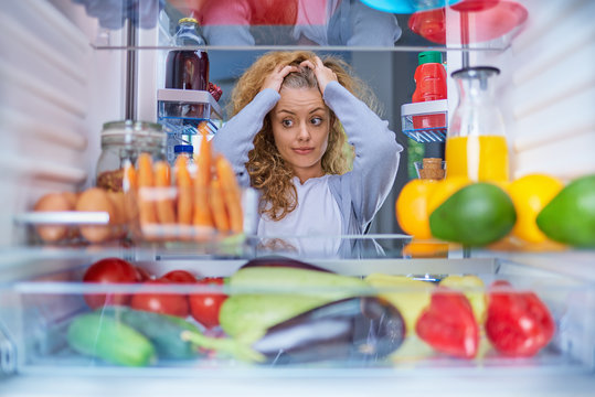 Confused Hungry Woman Holding Hands On Head While Standing In Front Of Opened Fridge Full Of Groceries.